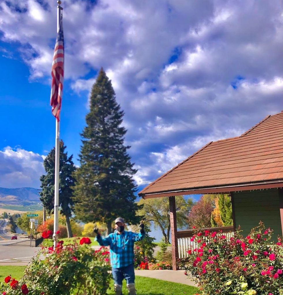 Exterior of a building with rose bushes and a flag pole flying the American flag. In front of the rose bushes, a man stands. Mr. Cortez is the key to helping Lakeside achieve the best landscaping.